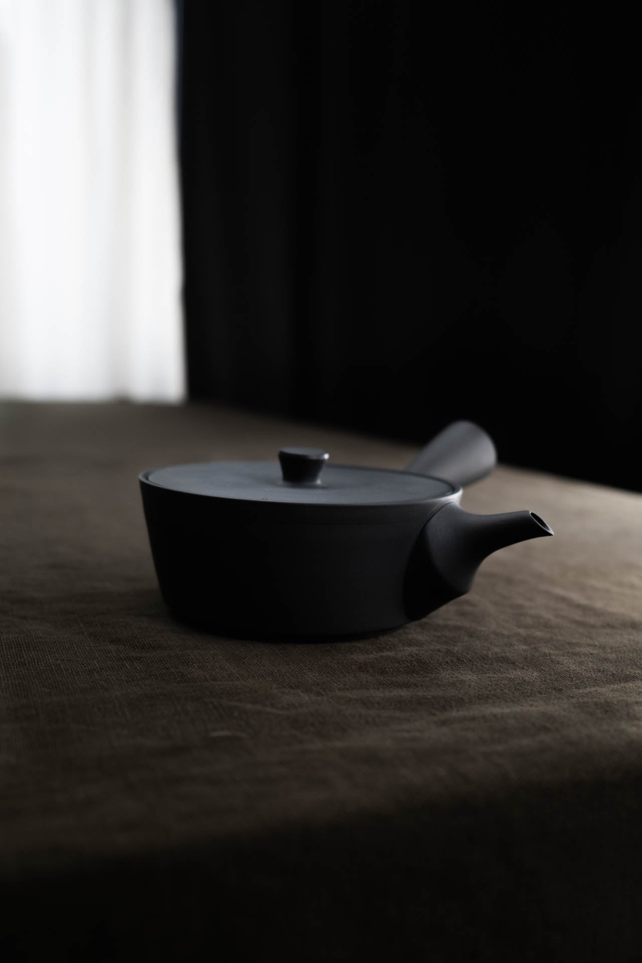 Kyusu Teekanne seitlich auf einem Tisch mit sandfarbenem Leinentischtuch, dunkler Hintergrund und weißem Teilhintergrund.